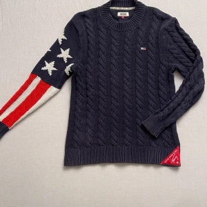 Mörkblå stickad Cable knit tröja från Tommy Jeans - Säljer en snygg mörkblå stickad tröja från Tommy Jeans. Den har ett amerikanskt flaggmönster på ena ärmen med stjärnor och ränder i vitt och rött. Tröjan är i mycket bra skick och perfekt för höst och vinter. Den har en klassisk kabelstickad design och rund halsringning. 
