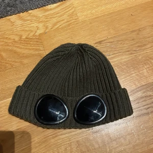 Olivgrön mössa från C.P. Company - Säljer en cool olivgrön mössa från C.P. Company. Den är stickad i 100% ull och har de ikoniska glasögon-detaljerna framtill. Perfekt för att hålla sig varm och stilren under höst och vinter. Tillverkad i Italien, vilket garanterar hög kvalitet. Passar perfekt för dig som vill sticka ut!
