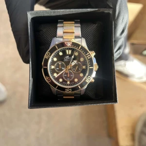 Svart och guld klocka från Lotus - Snygg Lotus-klocka i svart och guld med robust design. Den har en svart urtavla med guldiga detaljer och en tvåfärgad länkarmband i silver och guld. Perfekt för både vardag och fest!