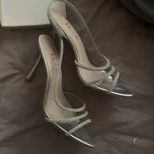 Silvriga klackskor med strassdetaljer - Snygga silvriga klackskor med glittrande strassdetaljer. De har en elegant design med smala remmar och en spetsig tå. Perfekta för festliga tillfällen när du vill glänsa lite extra. Klackarna är höga och ger en stilren look. Köpte utomlands för 700, säljer för 400 kan gå ner i pris. Aldrig använda pågrund av storlek 