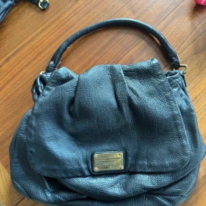 Svart skinnväska från Marc by Marc Jacobs - Säljer en snygg svart skinnväska från Marc by Marc Jacobs. Väskan har en stilren design med en metallplatta framtill och ett mönstrat foder. Insidan är tvättad men kommer inte bli vitare än på bild, men har tydliga spår av användning. Nött på metall och handtag men har mycket kvar att ge. Cirka 40 bred och 25 hög. 