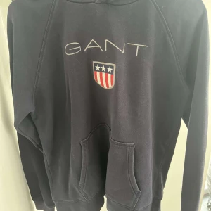 Svart hoodie från GANT - Säljer en snygg svart hoodie från GANT med deras ikoniska logga och ett coolt emblem på bröstet. Perfekt för både vardag och chill. Den är i bra skick och superbekväm att ha på sig. Passar perfekt för höst och vinter!