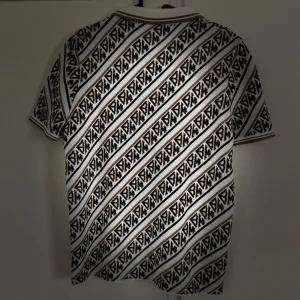 Mönstrad pikétröja med dragkedja - Säljer en snygg pikétröja med ett unikt diagonalt mönster i svart och beige. Tröjan har en dragkedja framtill och korta ärmar, perfekt för en stilren look. Passar både till vardags och festliga tillfällen!