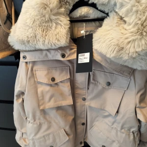 Beige parkajacka med fuskpäls - Säljer en superfin beige parkajacka från K-Zell. Den har en stor och mysig fuskpäls på huvan och praktiska fickor med knappar framtill. Perfekt för kyliga höst- och vinterdagar. Jackan är i nyskick och har en dragkedja samt knappar för extra skydd mot kylan.