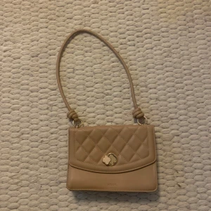 Beige quiltad axelväska från Fossil - Säljer en superfin beige axelväska från Fossil. Den har ett quiltat mönster och en snygg gulddetalj på framsidan. Väskan är i syntetmaterial och har en praktisk dragkedja inuti. Perfekt för både vardag och fest!