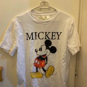 Vit t-shirt med Mickey Mouse från H&M - Säljer en vit t-shirt från H&M med en stor tryck av Mickey Mouse på framsidan. T-shirten är kortärmad och i bomull, perfekt för vardagsbruk eller sommarhäng. Den är i bra skick och har en normal passform. Disney-fans, missa inte denna! 🐭
