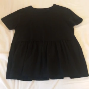 Svart kortärmad topp i storlek S - Säljer en svart kortärmad t-shirt/topp i storlek S. Den har en lös passform med en volangdetalj nedtill som ger en fin siluett. Perfekt för både vardag och festliga tillfällen. Den är ganska stor i  storleken. T-shirten är köps secondhand och lappen var bortklippt när jag köpte den så vet tyvärr inte vilket märke den är ifrån.💗