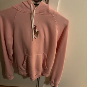 Rosa hoodie från Ralph Lauren - Säljer en superfin rosa hoodie från Ralph Lauren med den klassiska broderade polospelaren på bröstet. Den är i mjukt material och har en stor ficka fram samt justerbar huva med vita snören. Perfekt för en avslappnad stil eller kyliga dagar. 🩷