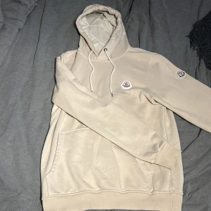 Beige hoodie från Moncler - Säljer en snygg beige hoodie från Moncler med  loga på bröstet även en klassisk loga på armen. Hoodien har en känguruficka och justerbar dragsko i luvan. Perfekt för en stilren och bekväm look. 