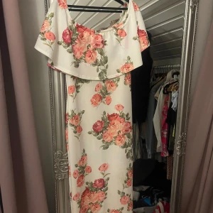 Blommig långklänning med volang - Säljer en romantisk långklänning med blommönster i rosa och grönt på vit botten. Klänningen har en offshoulder-design med volangdetalj som ger en feminin touch. Perfekt för sommarens festligheter!