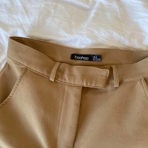 Beige byxor från Boohoo - Snygga beige byxor från Boohoo med hög midja och dragkedja i sidan. Byxorna har en stilren design och är perfekta för en chic look. De är tillverkade i ett mjukt material som ger en bekväm passform.
