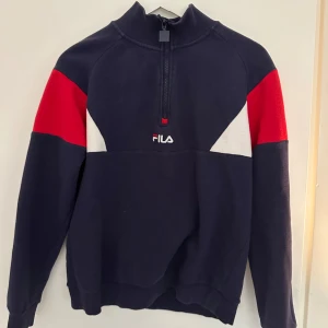 Mörkblå tröja med dragkedja från FILA - Säljer en snygg mörkblå tröja från FILA med dragkedja och hög krage. Tröjan har röda och vita detaljer på ärmarna och bröstet. Perfekt för en sportig look eller en avslappnad dag. Passar bra till jeans eller träningsbyxor.