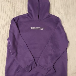 Lila hoodie med tryck - Säljer en lila hoodie med texten 'HOWEVER LONG THE NIGHT THE DAWN WILL BREAK' på bröstet. Baksidan har ett tryck av två händer. Perfekt för en avslappnad stil.