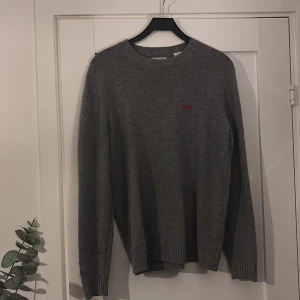 Grå stickad tröja från Levi's - Säljer en stilren grå stickad tröja från Levi's. Tröjan har en rund halsringning och långa ärmar, perfekt för kyligare dagar. Den har en liten röd logotyp på bröstet som ger en snygg detalj. Passar bra till både jeans och chinos.