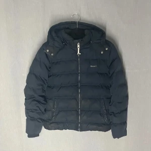 Svart pufferjacka från GANT - Säljer en svart pufferjacka från GANT med huva och dragkedja. Jackan har en stilren design med GANT-logga på bröstet och längs dragkedjan. Perfekt för kyliga dagar. Jackan har inga märken eller defekter. Extra knappar till jackan kommer med på köpet