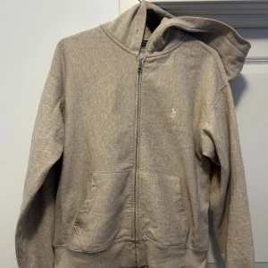 Beige hoodie från Ralph Lauren Small - Säljer en stilren beige hoodie från Ralph Lauren med dragkedja och huva. Tröjan har långa ärmar och en broderad logga på bröstet. Perfekt för en avslappnad look. Denna är i ett lite tunnare material.