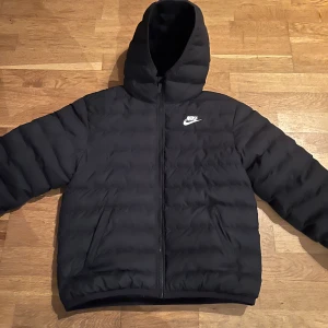 Nike jacka - Säljer en svart dunjacka från Nike med huva och dragkedja. Jackan har en klassisk pufferdesign och är perfekt för kyligare väder. Den har en broderad Nike-logga på bröstet och är tillverkad i Vietnam.