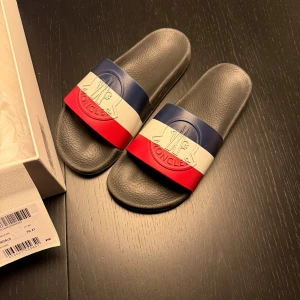 Moncler sandaler i blå, vit och röd - Snygga Moncler tofflor. Storlek 41 och i relativt fint skick. Likadana på farfetch och mintto ligger på cirka 2400kr