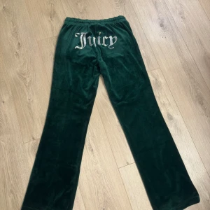 Gröna velourbyxor från Juicy Couture - Snygga gröna velourbyxor från Juicy Couture med glittrande logga på baksidan. Byxorna har en bekväm elastisk midja och dragkedjefickor fram. Perfekta för en avslappnad stil med en touch av glamour.