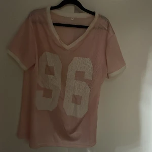 Rosa baseball/hockey meshtröja med nummertryck - Säljer en rosa baseball/hockey meshtröja med stort vitt nummertryck '96' på framsidan. Tröjan har en v-ringad hals och korta ärmar. Perfekt för en sportig look eller avslappnad stil. 🩷
