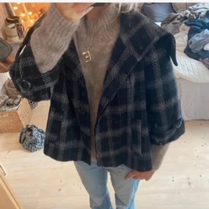 Kappa med trekvartsärm  - Snygg rutig kappa i svart och grått med stora knappar framtill. Den har en klassisk krage och trekvartsärmar, perfekt för höst och vår. En stilren och tidlös design som passar till många olika outfits.