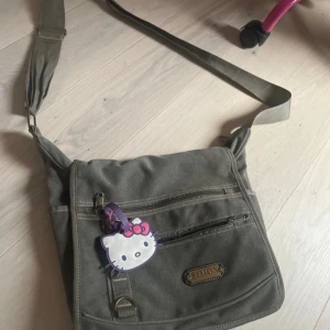 Grön axelväska från Simon - Säljer en grön axelväska från Simon med flera praktiska dragkedjefickor. Väskan har en justerbar axelrem och en söt Hello Kitty-nyckelring som detalj. Perfekt för att bära med sig det nödvändigaste på ett stilfullt sätt.