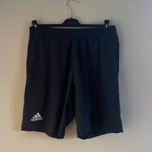 Mörkblå träningsshorts från Adidas - Snygga mörkblå träningsshorts från Adidas med blå detaljer. Perfekta för träning med bekväm passform och elastisk midja. Tillverkade av Primegreen-material för en hållbar känsla. Adidas-logga på benet ger en sportig look.