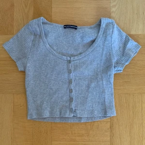 Grå ribbad croppad topp från Brandy Melville - Säljer en grå ribbad croppad topp från Brandy Melville. Toppen har korta ärmar och knappar framtill. Perfekt för en avslappnad stil. Storleken är okänd, men skulle gissa på S.