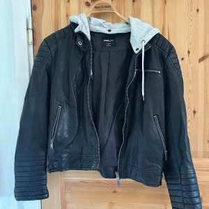 Svart skinnjacka med grå huva från FB Sister - Snygg svart skinnjacka från FB Sister med en avtagbar grå huva i sweatshirtmaterial. Jackan har dragkedjor på fickorna och ärmarna samt quiltade detaljer på axlarna och ärmsluten. Perfekt för en cool och avslappnad stil. Vid lappen på insidan så har det börjat lossna material men det är ingenting som syns när man har jackan på sig.