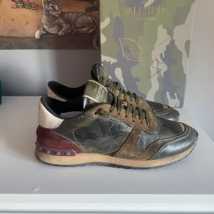 Valentino Rockrunner i camouflage - Snygga Valentino Rockrunner sneakers i ett coolt camouflage-mönster. Skorna har en kombination av gröna, svarta och vinröda nyanser med en vit häl. De är tillverkade i en mix av skinn och mocka och har snörning framtill. Perfekta för en stilren och trendig look.