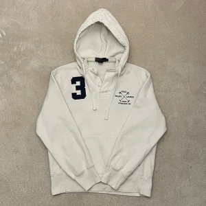 Vit hoodie från Ralph Lauren - Säljer en stilren vit hoodie från Ralph Lauren med en stor siffra '3' och broderad logga på bröstet. Tröjan har en klassisk känguruficka och knappar vid halsen. Perfekt för en avslappnad look. Vid minsta fråga eller fundering hör av er🤝