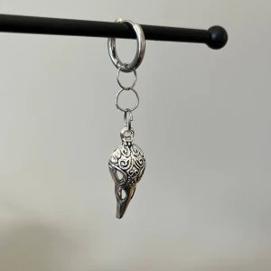 Silverfärgat örhänge i rostfritt stål med fågelhuvud - Unikt silverfärgat örhänge med ett detaljerat hänge i form av ett fågelhuvud. Hänget har ett intrikat mönster som ger en mystisk och elegant känsla. Perfekt för att ge din stil en unik touch.