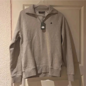 Khakifärgad zipup Morris - Säljer en stilren khakifärgad sweatshirt från Morris med halv dragkedja och en liten broderad detalj på bröstet. Perfekt för en avslappnad look. Tröjan har långa ärmar och ribbade muddar. Helt ny med lappar