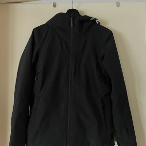Svart jacka från Peak Performance - Säljer en svart jacka från Peak Performance med text på huvan. Jackan är vattentät, vindtät och andas bra tack vare HIPE-materialet. Den har dragkedja och långa ärmar, perfekt för utomhusaktiviteter.