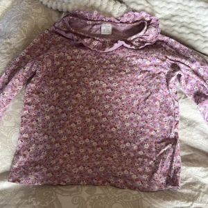 Lila blommig topp från Lindex - Söt lila topp från Lindex med blommigt mönster i vitt och gult. Toppen har långa ärmar och en volangdetalj vid halsringningen. Perfekt för en färgglad och lekfull stil!