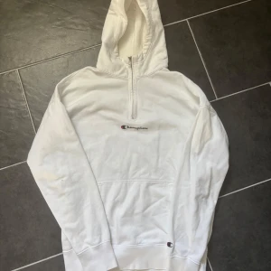 Vit hoodie från Champion - Säljer en vit hoodie från Champion med dragkedja vid halsen och klassisk logga på bröstet. Tröjan har en stor ficka framtill och långa ärmar. Baksidan av armbågen är skadad, men annars i väldigt bra skick