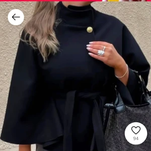 Missay poncho  - Elegant svart kappa med hög krage och dekorativ knappdetalj. Kappan har ett knytband i midjan för en justerbar passform och vida ärmar som ger en stilren look.