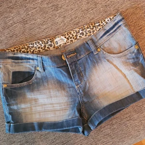 Blå jeansshorts - Snygga blå jeansshorts med leopardmönstrad insida. De har en klassisk femficksdesign och är lågmidjade med en knapp och dragkedja framtill. Perfekta för en avslappnad stil.