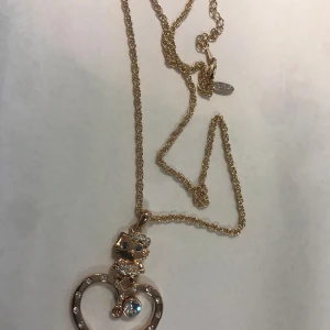 Hello Kitty Guldigt halsband med hjärtberlock - Charmigt guldigt halsband med en hjärtformad berlock. Berlocken har en söt figur och glittrande stenar som ger en extra touch av elegans. Perfekt för att lägga till lite gnista till din outfit.  Helt ny 