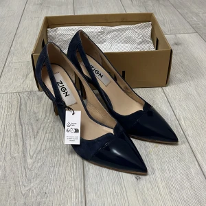 Svarta pumps från Zign - Eleganta marinblåa klackar från Zign med spetsig tå och hög klack köpt från zalando. Skorna har en stilren design med en kombination av glansigt och matt material. Perfekta för att ge en sofistikerad touch till din outfit. Kartong och mall finns kvar då de är helt nya! 