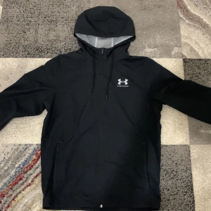 Svart Windbreaker från Under Armour - Säljer en svart vindjacka från Under Armour med huva och dragkedja. Jackan har en stilren design med logotyp på bröstet och är perfekt för blåsiga dagar. Den har en lös passform och är tillverkad i syntetmaterial för att stå emot väder och vind.