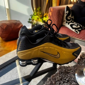Nike shox r4 i svart och guld - Snygga Nike sneakers i svart och guld med en futuristisk design. Skorna har en unik sula med synliga dämpningskuddar och klassisk snörning. Perfekta för dig som vill ha en sportig och stilren look.