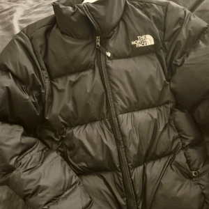 Svart dunjacka från The North Face - Säljer en svart dunjacka från The North Face. Jackan har en dragkedja framtill och är långärmad. Perfekt för kyliga dagar med sin pufferdesign som håller värmen inne. Logotypen är synlig både fram och bak.