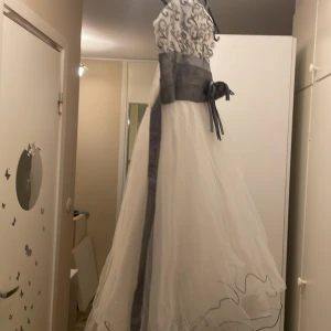 Vit och grå klänning med broderad överdel - Elegant långklänning i vitt med grå detaljer. Klänningen har en broderad överdel och ett brett grått band i midjan med en dekorativ rosett. Perfekt för formella tillfällen.