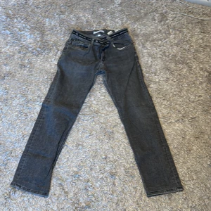 Grå jeans från Calvin Klein - Snygga grå jeans från Calvin Klein med klassisk femficksdesign och diskret CK-logga på linningen. Perfekta för en stilren look. De har en dragkedja och knapp framtill.