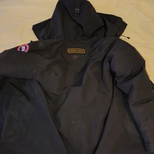 Svart dunjacka från Canada Goose - Säljer en svart dunjacka från Canada Goose med huva och dragkedja. Jackan har en broderad logotyp på ärmen och är perfekt för kalla dagar. Den är designad för att hålla dig varm och bekväm.