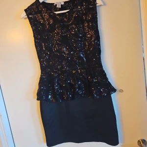 Svart glittrig klänning - Snygg svart klänning med glittriga paljetter och volangdetalj i midjan. Perfekt för en festlig kväll! Klänningen är ärmlös och har en elegant passform som framhäver figuren.