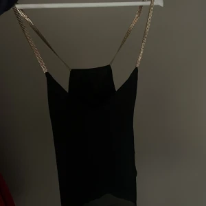 Svart linne med guldfärgade axelband - Elegant svart linne med smala guldfärgade axelband. Perfekt för en kväll ute eller en speciell tillställning. Linnet har en stilren design som framhäver en sofistikerad look.