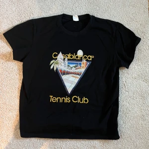 Svart t-shirt med Casablanca Tennis Club-tryck - Snygg svart t-shirt med ett färgglatt tryck av Casablanca Tennis Club. Perfekt för en avslappnad stil med en touch av retro. T-shirten har korta ärmar och en rund halsringning.