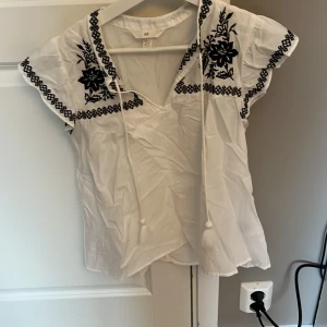 Vit blus med broderi från H&M - Säljer en vit blus från H&M med svart broderi och knytband framtill. Blusen har korta ärmar och ett vackert blommönster. Perfekt för en bohemisk look.